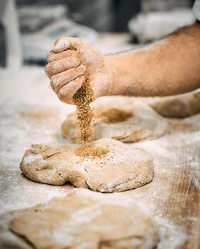 Hand von Bäckerei Schrott streut Körner über einen Teigling auf einer bemehlten Arbeitsfläche.