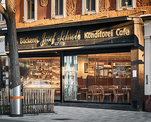 Außenansicht der Bäckerei Schrott mit Konditorei-Auslage, großer Fensterfront und Sitzplätzen im Café.