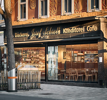 Außenansicht der Bäckerei Schrott mit großer Fensterfront, Konditorei-Auslage und gemütlichem Cafébereich.