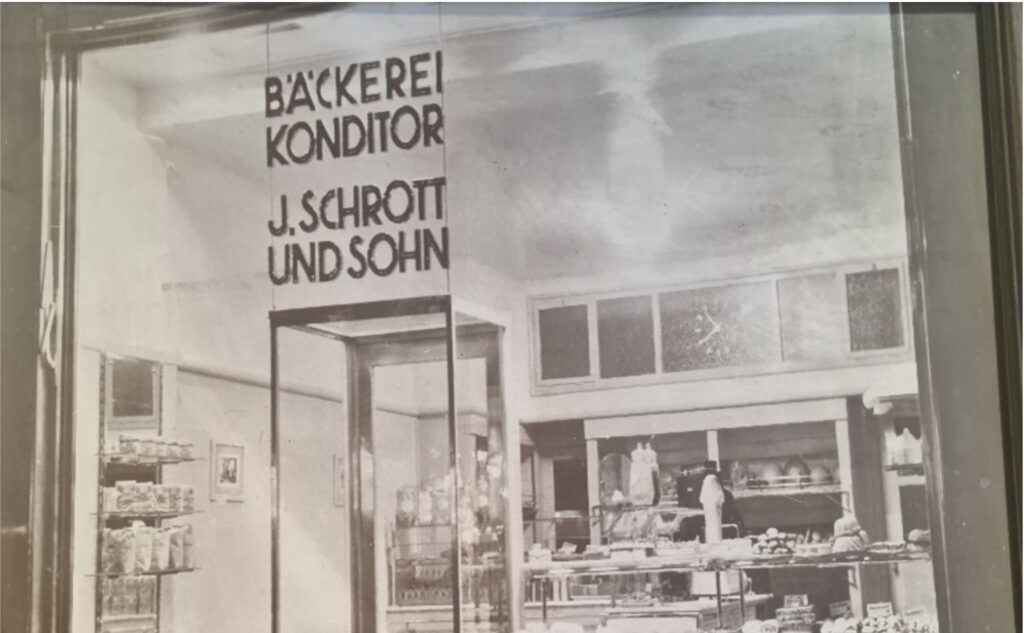 Historisches Foto der Verkaufsfiliale von Bäckerei Schrott mit Beschriftung „Bäckerei Konditor J. Schrott und Sohn“ und Blick in den Innenraum.