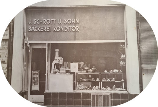 Historische Filiale von Bäckerei Schrott mit der Aufschrift „J. Schrott u. Sohn Bäckerei Konditor“ und Schaufenster mit Brot, Gebäck und Mehlspeisen.
