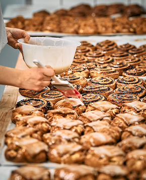 Hand von Bäckerei Schrott bestreicht frisch gebackene Zimtschnecken und Gebäck mit glänzender Glasur.